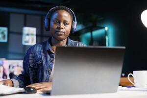 focado Preto mulher vestindo sem fio fones de ouvido usando computador portátil e escrevendo notas. africano americano fêmea aluna com fone de ouvido assistindo conectados webinário, ouvindo para audio curso em pessoal computador. foto