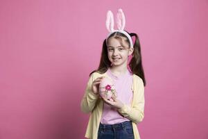 adorável fofa pequeno menina com Coelho orelhas mostra uma grande Rosa ovo, posando com Páscoa decorações e arranjos. jovem alegre criança ser saiu sobre Primavera celebração, feito à mão arte. foto