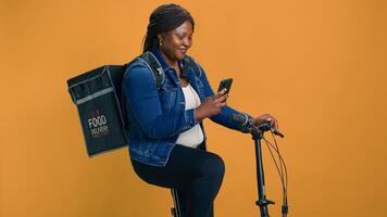 africano americano fêmea multitarefa em dela bicicleta, entregando Comida com celular dentro mão. jovem Preto mulher verificação Entrega aplicativo em Móvel dispositivo para eficiente transporte do ordens. foto