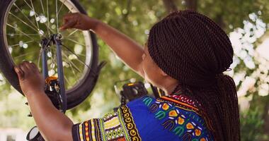 saudável Preto mulher consertando dela próprio bicicleta usando especializado equipamento a partir de kit de ferramentas dentro quintal. boneca mais Zoom tomada. fêmea africano americano ciclista reparação frente bicicleta roda ao ar livre. foto