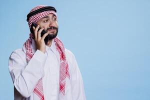 muçulmano homem vestido dentro islâmico roupas, respondendo ligar e tendo conversação em Móvel telefone. árabe pessoa vestido dentro tradicional Thobe e ghutra enquanto falando em Smartphone foto