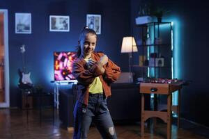 fofa menina realizando tendendo dança rotina dentro vivo sala, cercado de televisão tela exibindo 3d renderiza. pequeno criança dançando dentro rgb luzes aceso casa estúdio, criando conteúdo para conectados fãs foto