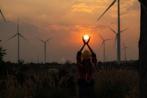 engenheiro mulher com amarelo capacete e vento turbina em pôr do sol fundo. foto