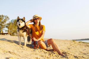 mulher dentro chapéu e oculos de sol senta com dela cachorro em a de praia e desfrutando pôr do sol foto