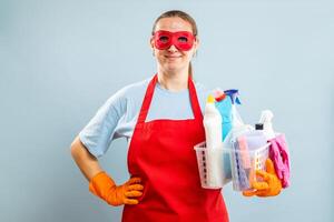 mulher dentro vermelho mascarar, luvas e avental segurando cesta com limpeza suprimentos foto