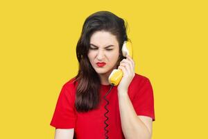 mulher com vermelho batom fala em a velho amarelo Telefone com uma cordão. mulher é infeliz, faz não quer para ouço para dela interlocutor, pega nervoso. indesejado ligar em uma amarelo fundo foto