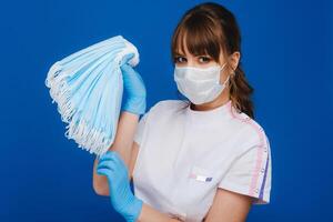 uma jovem menina médico dentro uma médico mascarar detém uma muitos do máscaras dentro dela mãos. uma enfermeira poses em uma azul fundo foto