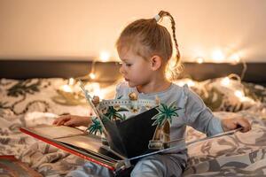 pequeno menina lendo pop acima livro dentro casa cama dentro Natal meio Ambiente com luzes fundo foto