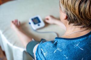 Senior mulher verificação sangue pressão nível às lar, Mais velho fêmea sofrimento a partir de Alto sangue pressão usando uma pulsômetro, tonômetro. costas Visão foto