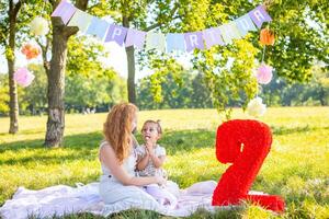 alegre mãe e filha tendo Diversão em criança aniversário em cobertor com papel decorações dentro a parque foto