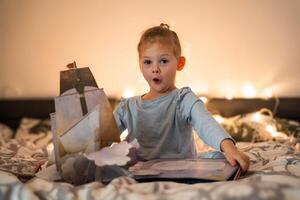 pequeno menina lendo pop acima livro dentro casa cama dentro Natal meio Ambiente com luzes fundo foto