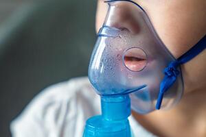 fechar acima Visão do pequeno menina estão sentado e segurando uma nebulizador mascarar inclinado contra a face, via aérea tratamento conceito foto