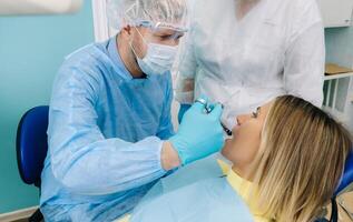 uma dentista vestindo uma protetora mascarar senta perto e detém instrumentos dentro dele mãos antes tratando uma paciente dentro a dental escritório foto