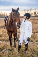 lindo loiro profissional fêmea jóquei em pé perto cavalo dentro campo dentro inverno. amizade com cavalo conceito foto