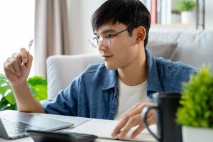 a homem dentro casual roupas trabalhando com uma computador portátil, computador, inteligente telefone, calculadora sentado em a sofá dentro a vivo quarto às lar, trabalhando a partir de casa conceito. foto