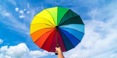mão segurando colorida arco Iris guarda-chuva e azul céu fundo. lgbt, orgulho mês, diversidade, protetor solar conceito. foto