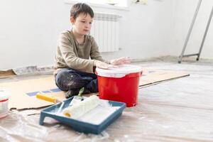 criança tem Diversão sentado em a chão e batendo dele mãos em uma balde do pintura foto