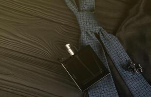 uma garrafa de colônia masculina e abotoaduras com gravata azul estão sobre um fundo de tecido preto de luxo em uma mesa de madeira. acessórios masculinos foto