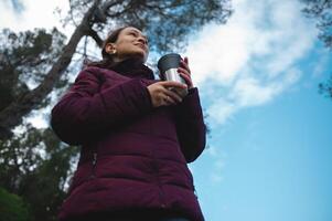 inferior visualizar. viajante mulher segurando uma garrafa térmica copo com quente bebida, chá ou café, olhando ausente, caminhada dentro a floresta foto