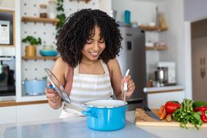 saudável Comida às lar. feliz mulher é preparando a apropriado refeição dentro a cozinha. lindo sorridente jovem mulher dentro avental em pé dentro cozinha e mexendo sopa dentro Panela. foto