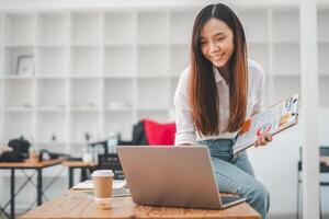 uma focado jovem mulher avaliações documentos enquanto trabalhando em dela computador portátil dentro uma brilhante e organizado escritório espaço. foto