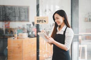 lindo jovem barista mulher dentro avental segurando ordem papel e em pé dentro frente do a porta do cafeteria com aberto placa borda. o negócio proprietário comece conceito. foto