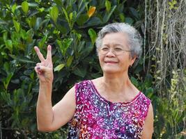 uma Senior ásia mulher com curto branco cabelo sorridente e olhando às a Câmera enquanto em pé dentro uma jardim foto