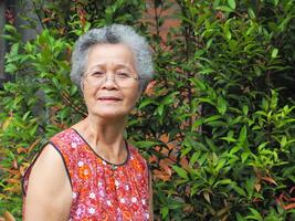 Senior ásia mulher com curto branco cabelo sorridente e olhando às a Câmera enquanto em pé dentro uma jardim. espaço para texto. conceito do envelhecido pessoas e cuidados de saúde foto