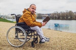 paraplégico deficiente homem dentro cadeira de rodas é lendo livro ar livre. foto