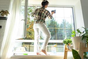 mulher manualmente lavagens a janela do a casa com uma trapo com spray limpador e esfregão dentro a interior com branco cortinas. restaurando ordem e limpeza dentro a primavera, limpeza servise foto