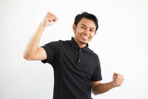 sorridente ou feliz jovem ásia homem cerrado dele punhos torcendo despreocupado e excitado. vitória conceito, vestindo Preto pólo t camisa isolado em branco fundo foto