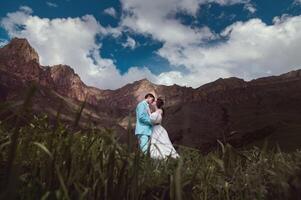 uma jovem homem e dele esposa ficar de pé dentro a abraço Alto dentro a montanhas contra a pano de fundo do épico pedras em uma ensolarado dia. recém-casados Casamento casal dentro a montanhas foto