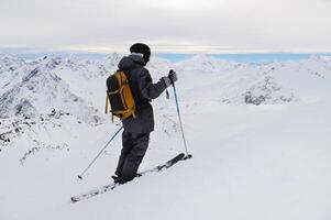 lado Visão do a Atlético esquiador dentro uma capacete e óculos com uma mochila, em pé em esquis, segurando esqui postes dentro branco neve, contra a céu, desfrutando uma lindo Visão do a montanhas foto