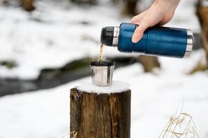 masculino mão derramando quente chá a partir de uma garrafa térmica para dentro uma caneca, fechar-se. quente beber com vapor dentro inverno. inverno caminhada conceito, acampamento, viagem. foto