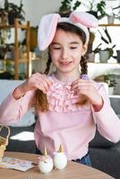 uma fofa menina com Rosa Coelho orelhas faz a Páscoa construir - decora a ovo dentro a Formato do uma unicórnio com strass, buzina, flores dentro a interior do uma casa com plantas. foto