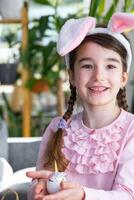 uma fofa menina com Rosa Coelho orelhas faz a Páscoa construir - decora a ovo dentro a Formato do uma unicórnio com strass, buzina, flores dentro a interior do uma casa com plantas. foto