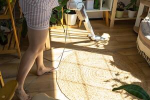 uma mulher aspiradores uma volta tapete dentro uma casa entre casa plantas com uma mão vácuo limpador. geral limpeza do a casa, limpeza serviço e dona de casa foto