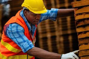 africano trabalhador carpinteiro vestindo segurança uniforme e Difícil chapéu trabalhando e verificação a qualidade do de madeira produtos às oficina fabricação. homem e mulher trabalhadores madeira dentro Sombrio armazém indústria. foto