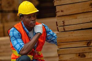 africano trabalhador carpinteiro vestindo segurança uniforme e Difícil chapéu trabalhando e verificação a qualidade do de madeira produtos às oficina fabricação. homem e mulher trabalhadores madeira dentro Sombrio armazém indústria. foto