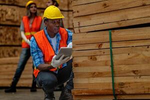 africano trabalhador carpinteiro vestindo segurança uniforme e Difícil chapéu trabalhando e verificação a qualidade do de madeira produtos às oficina fabricação. homem e mulher trabalhadores madeira dentro Sombrio armazém indústria. foto