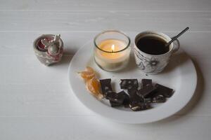 uma vela, copo do café e Preto chocolate em a branco placa. foto