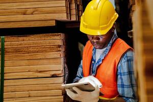 africano trabalhador carpinteiro vestindo segurança uniforme e Difícil chapéu trabalhando e verificação a qualidade do de madeira produtos às oficina fabricação. homem e mulher trabalhadores madeira dentro Sombrio armazém indústria. foto