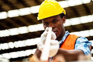 africano trabalhador carpinteiro vestindo segurança uniforme e Difícil chapéu trabalhando e verificação a qualidade do de madeira produtos às oficina fabricação. homem e mulher trabalhadores madeira dentro Sombrio armazém indústria. foto