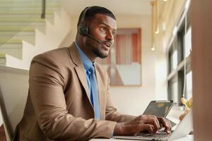 cliente Apoio, suporte ligar Centro operador ou recepcionista. cliente serviço e homem consultor falando enquanto fazendo uma consulta on-line. africano operador masculino telemarketing agente em uma computador portátil dentro escritório foto