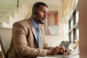 cliente Apoio, suporte ligar Centro operador ou recepcionista. cliente serviço e homem consultor falando enquanto fazendo uma consulta on-line. africano operador masculino telemarketing agente em uma computador portátil dentro escritório foto