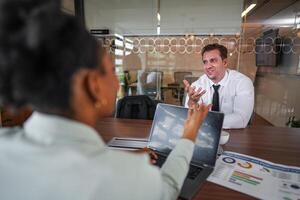 atraente africano jovem confiante empresária sentado às a escritório mesa com grupo do colegas dentro a fundo. trabalhando em computador portátil computador. bem sucedido mulher empreendedor. entrevista Novo funcionários. foto