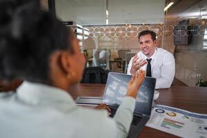 atraente africano jovem confiante empresária sentado às a escritório mesa com grupo do colegas dentro a fundo. trabalhando em computador portátil computador. bem sucedido mulher empreendedor. entrevista Novo funcionários. foto