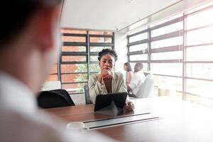 atraente africano jovem confiante empresária sentado às a escritório mesa com grupo do colegas dentro a fundo. trabalhando em computador portátil computador. bem sucedido mulher empreendedor. entrevista Novo funcionários. foto