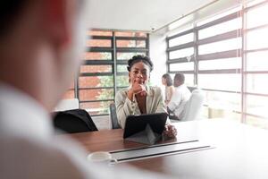 atraente africano jovem confiante empresária sentado às a escritório mesa com grupo do colegas dentro a fundo. trabalhando em computador portátil computador. bem sucedido mulher empreendedor. entrevista Novo funcionários. foto