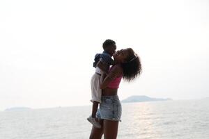 misturado raça africano e ásia mãe e Garoto é jogando às a ao ar livre área. sorridente feliz família ter Diversão corrida em a de praia. retrato do mãe e criança estilo de vida com uma único Penteado. foto